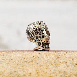 Puede incluir: Un anillo de calavera de plata con piedras negras engastadas en la cara de la calavera. El anillo est&aacute; decorado con dise&ntilde;os intrincados y est&aacute; sobre una superficie marr&oacute;n claro.