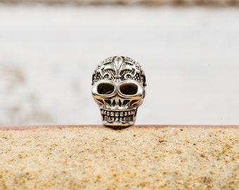 Puede incluir: Una detallada cuenta de calavera plateada con intrincados diseños florales y de flor de lis. La cuenta tiene cuencas de ojos grandes y una mandíbula detallada. La calavera es un elemento decorativo, posiblemente para joyería u otras manualidades.