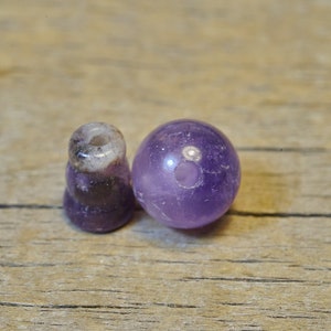 May include: Two amethyst gemstone beads, one round and one cylindrical, with a hole drilled through the center of each. The beads are a light purple color with a slight shimmer.