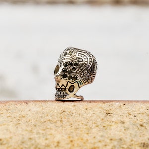 Puede incluir: Un anillo de calavera de plata con intrincados detalles en blanco y negro. La calavera est&aacute; adornada con peque&ntilde;as piedras negras.