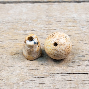 May include: Two natural stone beads with holes drilled through the center. The beads are a light brown color with darker brown and black speckles. The beads are sitting on a wooden surface.