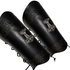 May include: A pair of black leather arm guards with a silver Mjolnir hammer design. The guards are laced up the side and have a Celtic knot pattern.