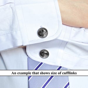 May include: A pair of silver cufflinks with a moon design. The cufflinks are shown on a white dress shirt with a blue and white striped tie.