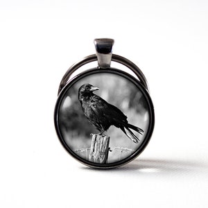 May include: Black and white photograph of a crow perched on a wooden fence post. The photo is on a round, silver keyring.
