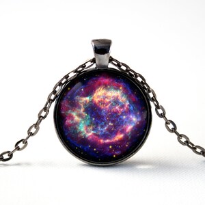 Puede incluir: Un collar de cadena plateada con un colgante redondo de vidrio que presenta una imagen de nebulosa colorida. La nebulosa es una mezcla de colores rosa, morado, azul y amarillo.