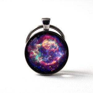 Puede incluir: Un llavero plateado con un charm redondo de vidrio que presenta una imagen de nebulosa colorida. La nebulosa es una mezcla de colores rosa, morado, azul y naranja con una dispersi&oacute;n de estrellas.