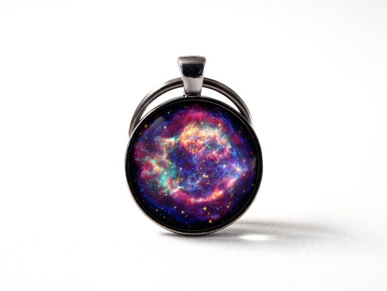 Puede incluir: Un llavero plateado con un charm redondo de vidrio que presenta una imagen de nebulosa colorida. La nebulosa es una mezcla de colores rosa, morado, azul y naranja con una dispersi&oacute;n de estrellas.