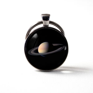 Peut inclure: Un porte-clés argenté avec un charme rond en verre représentant une image détaillée de Saturne avec ses anneaux sur un fond noir.