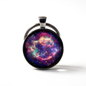 May include: A silver key chain with a round glass charm featuring a colorful nebula image. The nebula is a swirling mix of pink, purple, blue, and orange with a scattering of stars.