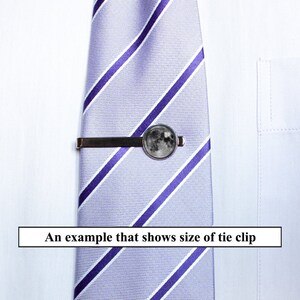 May include: A silver tie clip with a round moon-shaped design. The tie clip is attached to a striped purple and white tie.