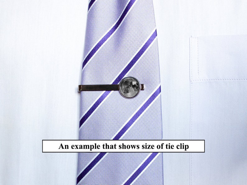 May include: A silver tie clip with a round moon-shaped design. The tie clip is attached to a striped purple and white tie.