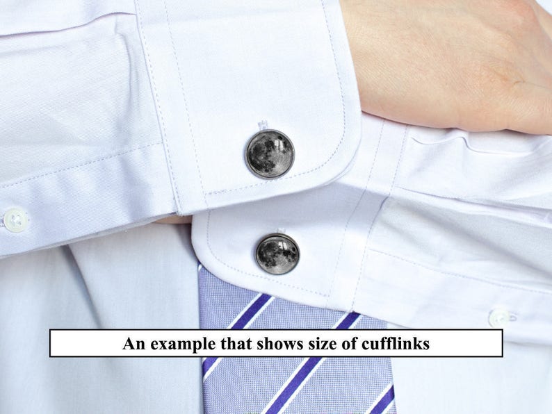 May include: Close-up of a white dress shirt with two cufflinks. The cufflinks are round with a detailed image of the moon. A blue and white striped tie is visible. The text in the image reads: "An example that shows size of cufflinks."