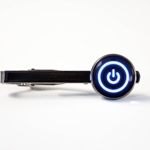 Puede incluir: Un pasador de corbata negro con un cabujón de vidrio redondo con borde plateado que presenta un símbolo de botón de encendido blanco sobre un fondo negro.