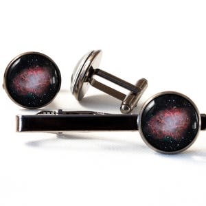 Op de afbeelding: Een set manchetknopen en een stropdasclip met een rood en zwart nevelontwerp. De manchetknopen zijn zilverkleurig met een ronde, zwarte achtergrond en de stropdasclip is zilverkleurig met een zwarte achtergrond.