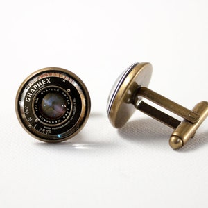 May include: A pair of cufflinks featuring a vintage camera lens with the text "GRAPHEX" and "OPTAR" printed on the lens. The cufflinks are made of brass and have a silver finish.