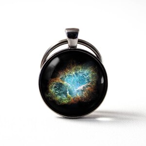 May include: A silver key chain with a round glass charm featuring a colourful nebula image. The nebula is a swirling mix of blue, green, yellow, and orange.
