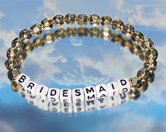 May include: A gold beaded bracelet with white letter beads spelling out the word "BRIDESMAID".