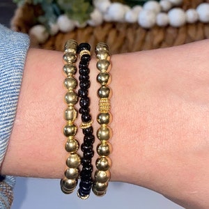 May include: Three bracelets stacked on a wrist. The bottom bracelet is made of gold beads, the middle bracelet is made of black beads, and the top bracelet is made of gold beads with a gold band.