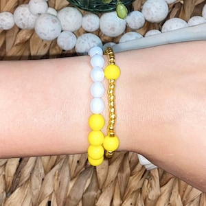 Puede incluir: Dos pulseras de cuentas, una blanca y otra amarilla, puestas en una muñeca.