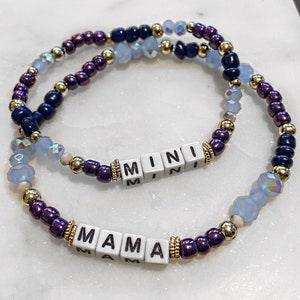 May include: Two beaded bracelets with the words "MAMA" and "MINI" spelled out in white letter beads. The bracelets are made with gold, purple, and blue beads.