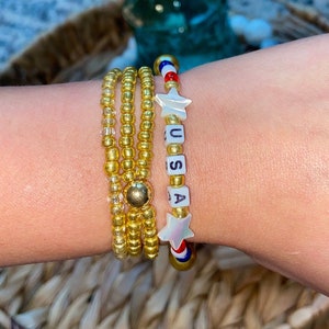 Puede incluir: Un brazalete de cuentas doradas con un dije de estrella rojo, blanco y azul y la palabra "USA" escrita en cuentas blancas.