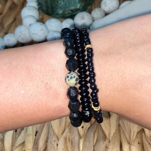 Puede incluir: Un conjunto de tres pulseras con cuentas negras y una sola cuenta más grande y moteada. Las pulseras están apiladas en la muñeca de una persona.