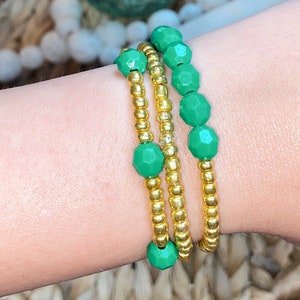 Puede incluir: Tres pulseras apiladas en una muñeca. Las pulseras están hechas de pequeñas cuentas doradas y cuentas verdes facetadas más grandes.