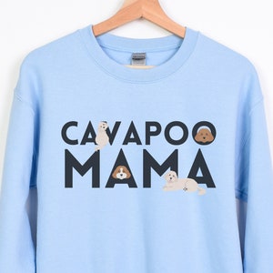 Op de afbeelding: Lichtblauw sweatshirt met de tekst "CAVAPOO MAMA" in zwarte blokletters. Boven het woord "CAVAPOO" staan drie kleine hondenillustraties. Het sweatshirt hangt aan een houten hanger. De woorden "LIGHT BLUE" staan in een cirkel onderaan.