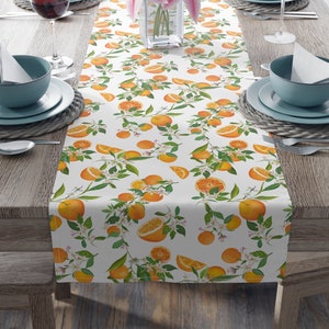 May include: White table runner with a repeating pattern of orange slices and green leaves. The runner is on a wooden table with two place settings on each side.