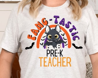 May include: A light gray t-shirt with a Halloween-themed design. The shirt features the words "FANG-TASTIC" and "PRE-K TEACHER" in orange and purple, along with a cartoon bat and rainbow graphic. Black bat silhouettes are scattered around the design.