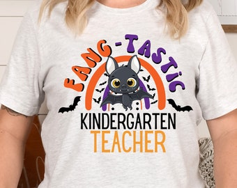May include: Light gray t-shirt with a Halloween-themed graphic. The design features a cartoon bat with the text "FANG-TASTIC KINDERGARTEN TEACHER" in orange, purple, and black. The shirt has a crew neck and short sleeves.