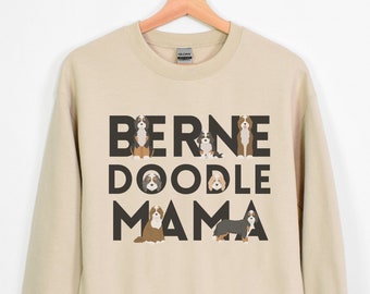 Puede incluir: Una sudadera de color arena con el texto "BERNE DOODLE MAMA" en letras negras. Las letras están rodeadas de ilustraciones de perros de montaña de Berna.