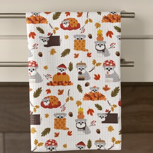 Puede incluir: Toalla de cocina blanca con un patrón repetido de perros grises, hojas de otoño, setas y calabazas. La toalla está colgada de una barra plateada. El diseño presenta colores otoñales.