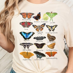 May include: A cream-colored t-shirt with a graphic of various butterfly species, including the Monarch Butterfly, Red Admiral, Clouded Sulphur, Loner Moth, Peacock, Painted Lady Butterfly, Canadian Tiger Swallowtail, Blue Morpho, Zebra Butterfly, Giant Owl Butterfly, Giant Swallowtail, Mourning Cloak, Great Spangled Fritillary, Variegated Fritillary, Spicebush Swallowtail, and Question Mark Butterfly.
