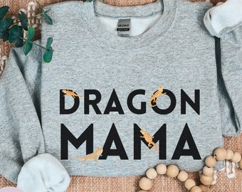May include: A grey sweatshirt with the text "Dragon Mama" in black lettering. The sweatshirt is decorated with small, orange, lizard-like figures.