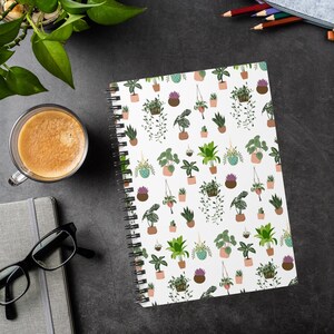 May include: A spiral-bound notebook with a white cover featuring a repeating pattern of potted plants in various sizes and styles. The plants are in shades of green, brown, and purple. The notebook is open, revealing blank pages.