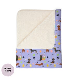 May include: A purple and white sherpa fleece throw blanket with a pattern of dachshunds, Easter eggs, and paw prints. The blanket is folded in half, showing the sherpa fleece side on top and the printed side underneath.