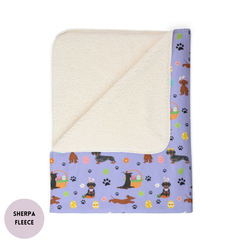 May include: A purple and white sherpa fleece throw blanket with a pattern of dachshunds, Easter eggs, and paw prints. The blanket is folded in half, showing the sherpa fleece side on top and the printed side underneath.