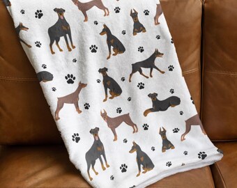 Puede incluir: Manta de forro polar blanca con un patrón repetido de Doberman Pinschers marrones y negros y huellas de patas.
