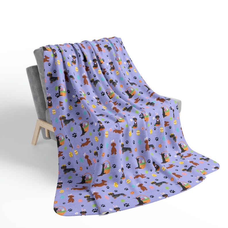 May include: A purple fleece throw blanket with a repeating pattern of dachshund dogs holding Easter baskets filled with colorful eggs. The dogs are brown and black with long ears and short legs. The background is a light purple with paw prints and small flowers.