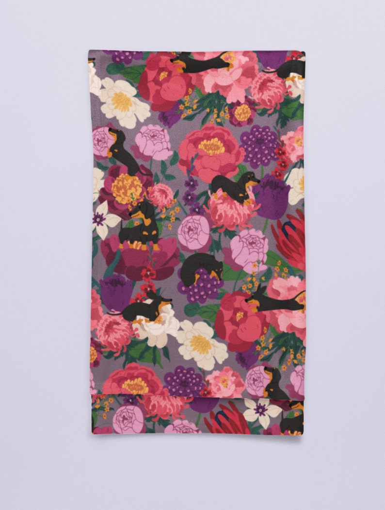 May include: A purple fabric tea towel with a floral pattern featuring black and tan dachshund dogs. The flowers are pink, red, white, and yellow.