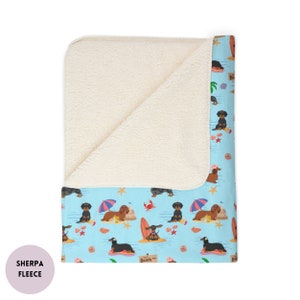 May include: A soft, white sherpa fleece blanket with a blue and white printed underside featuring a pattern of dachshunds on the beach. The pattern includes dachshunds surfing, building sandcastles, and relaxing under umbrellas. The text "Beach" is visible on the blanket.