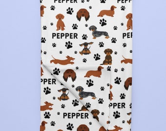 Puede incluir: Manta blanca con un patrón repetido de ilustraciones de perros salchicha en varias poses y colores, incluyendo marrón, negro y gris. También aparecen huellas de patas negras y la palabra "PEPPER".
