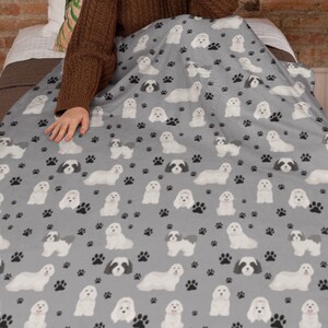K&ouml;nnte beinhalten: Eine graue Fleece-Decke mit einem sich wiederholenden Muster aus wei&szlig;en Malteser-Hunden und schwarzen Pfotenabdr&uuml;cken.