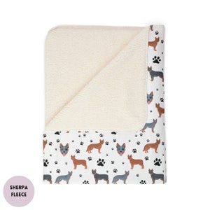 Peut inclure: Une couverture en polaire sherpa blanche avec un imprim&eacute; blanc et noir de chiens bergers australiens et d'empreintes de pattes. La couverture est pli&eacute;e en deux, montrant la polaire sherpa d'un c&ocirc;t&eacute; et le c&ocirc;t&eacute; imprim&eacute; de l'autre.
