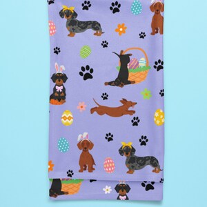May include: Purple tea towel with a repeating pattern of dachshund dogs wearing bunny ears and holding Easter eggs. The dogs are in various poses, including sitting, standing, and running. There are also paw prints and flowers on the towel.