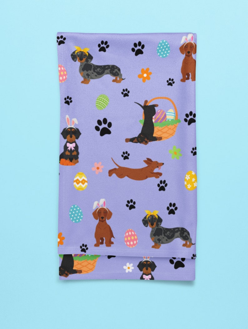 May include: Purple tea towel with a repeating pattern of dachshund dogs wearing bunny ears and holding Easter eggs. The dogs are in various poses, including sitting, standing, and running. There are also paw prints and flowers on the towel.