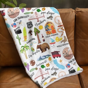 May include: White beach towel featuring a colourful pattern of Californian landmarks, including the Golden Gate Bridge, palm trees, a surfboard, and the words "San Francisco", "Los Angeles", "Sacramento", "San Diego", "Anaheim", "Palm Springs", "Hollywood", and "Oakland".