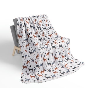 Peut inclure: Une couverture en polaire blanche avec un motif r&eacute;p&eacute;t&eacute; de silhouettes de chiens de b&eacute;tail australiens et d'empreintes de pattes. La couverture est drap&eacute; sur un fauteuil gris.