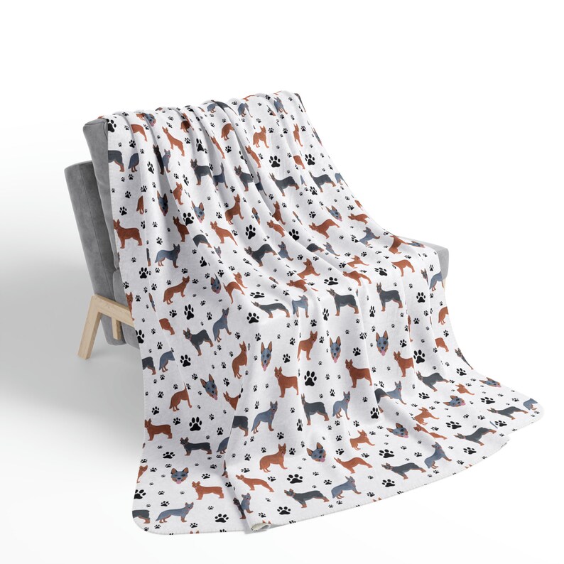 Peut inclure: Une couverture en polaire blanche avec un motif r&eacute;p&eacute;t&eacute; de silhouettes de chiens de b&eacute;tail australiens et d'empreintes de pattes. La couverture est drap&eacute; sur un fauteuil gris.